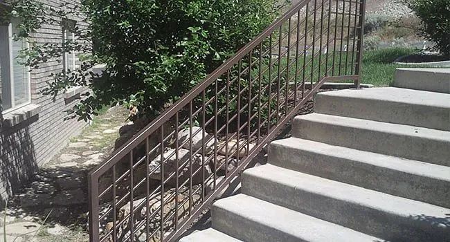 Concrete outdoor stairs with a brown metal railing beside a building and leafy greenery.