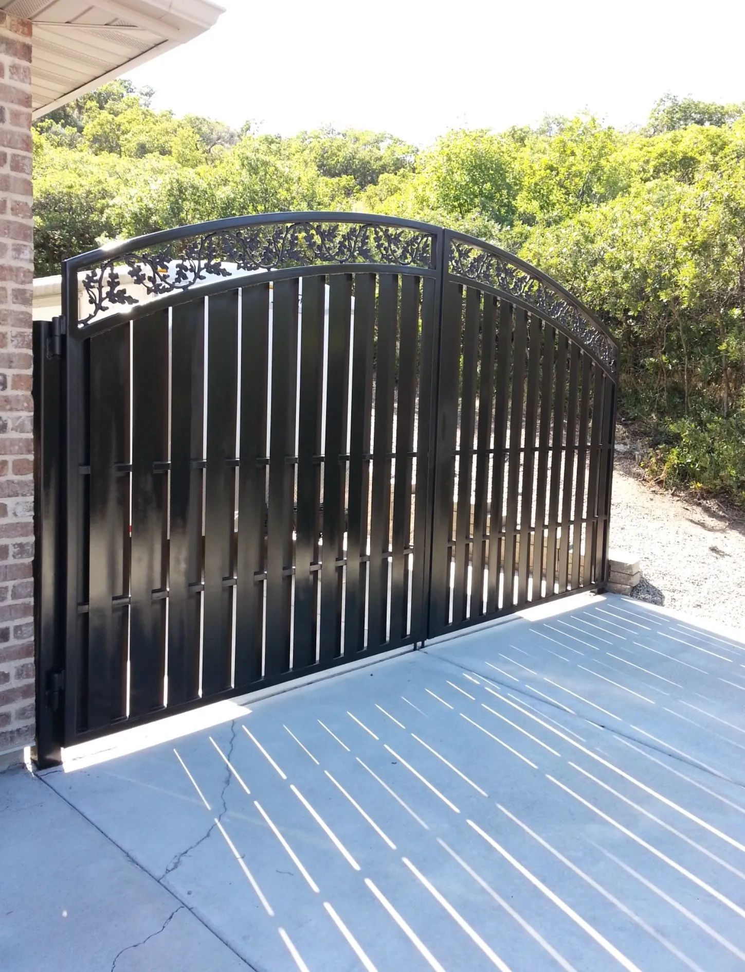 Black metal driveway gate beside a brick wall, with sunlit blue deck and green shrubs behind it