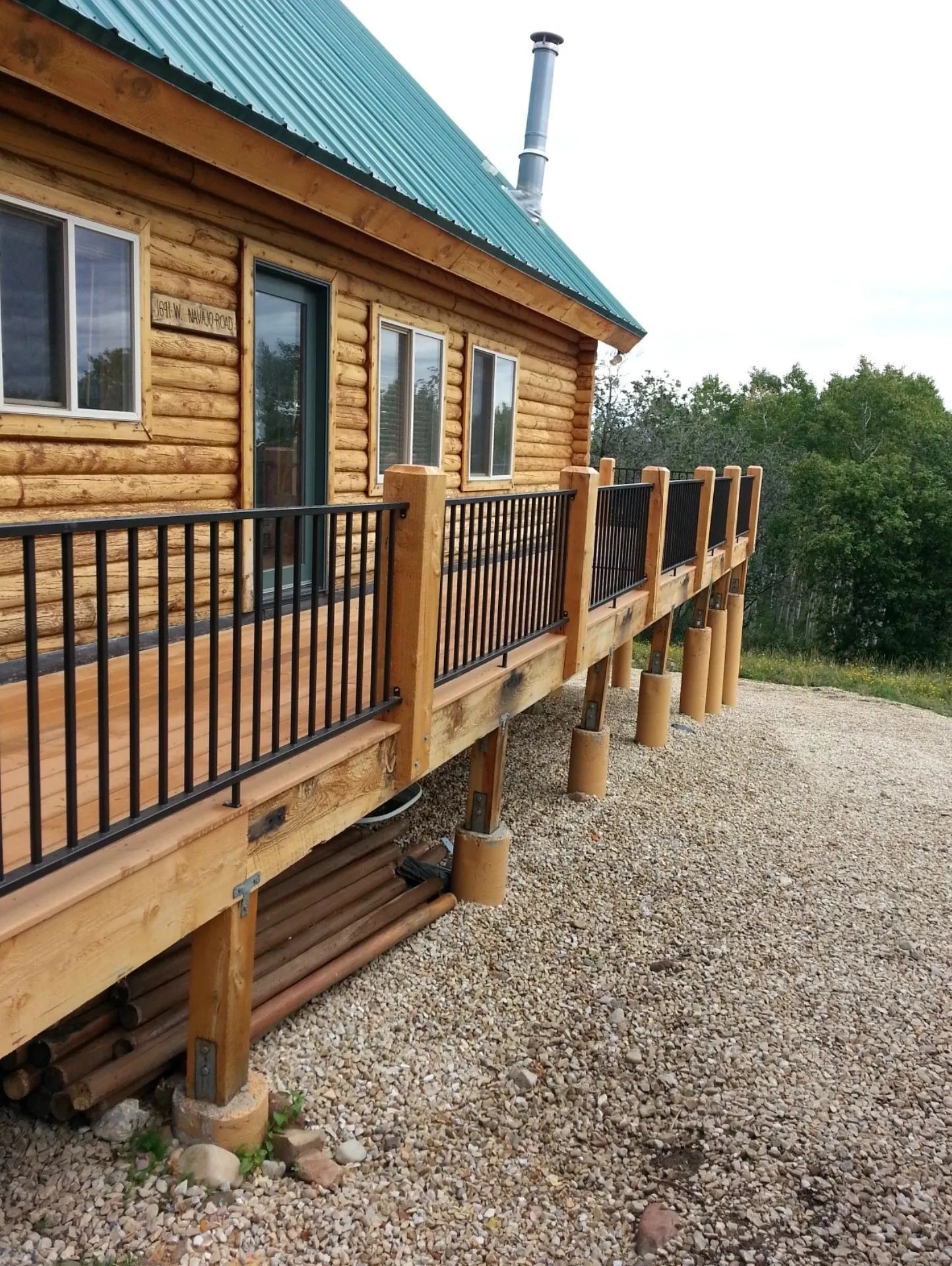 Wooden cabin with a raised deck and railing beside a gravel driveway, surrounded by trees.