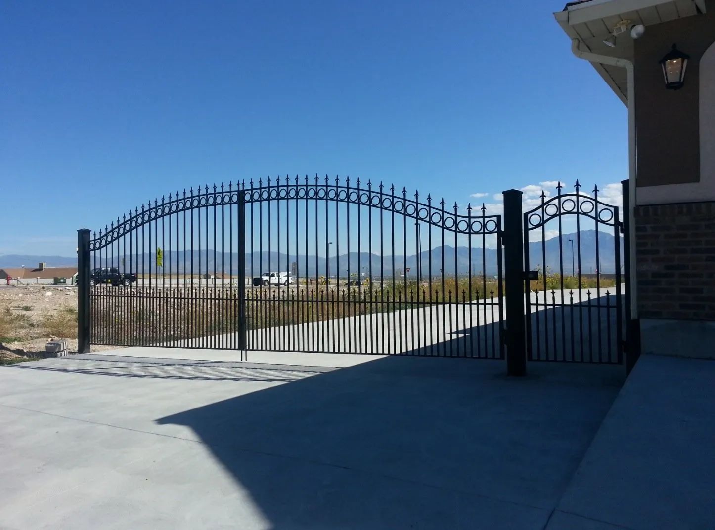 Black wrought-iron gate beside a house on a concrete driveway under a clear blue sky