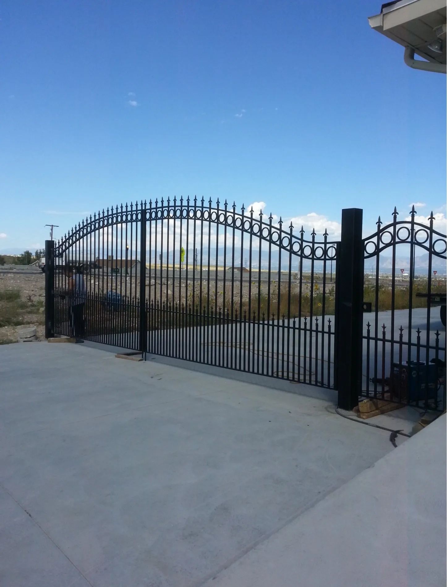 Black wrought-iron driveway gate beside a concrete driveway under a clear blue sky
