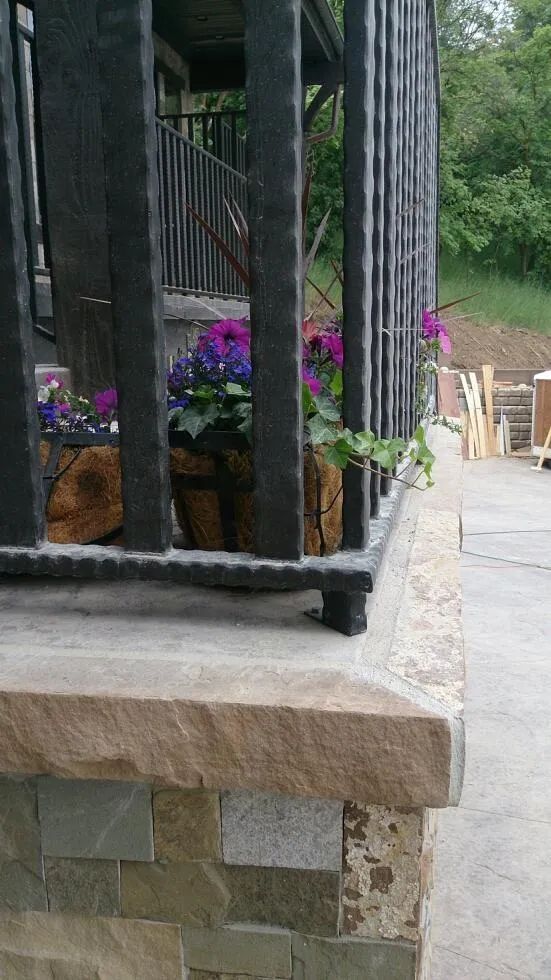 Stone porch with black railing and purple potted flowers beside a walkway and chairs outdoors