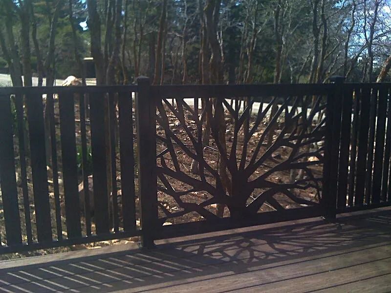Black metal gate with a tree-branch design, casting shadows on a wooden deck in a wooded area.