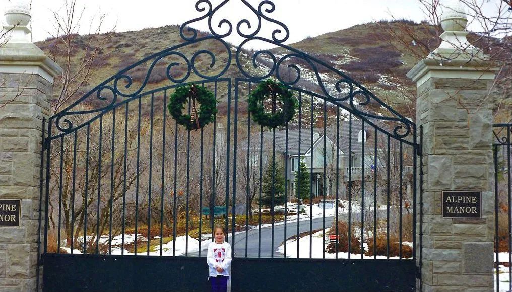 Ornate black wrought-iron gate with holiday wreaths, stone pillars, and a person standing in front.