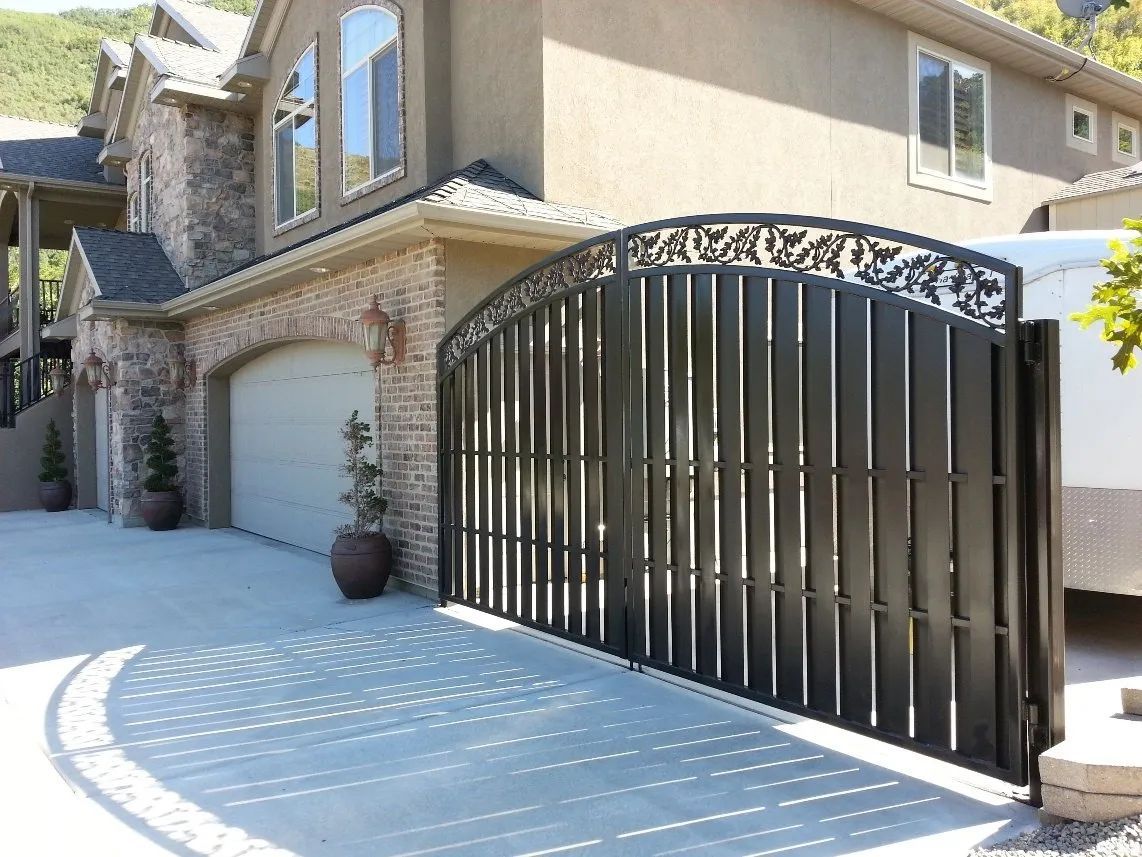 Black metal driveway gate beside a stone-and-stucco house with a garage and curved concrete driveway