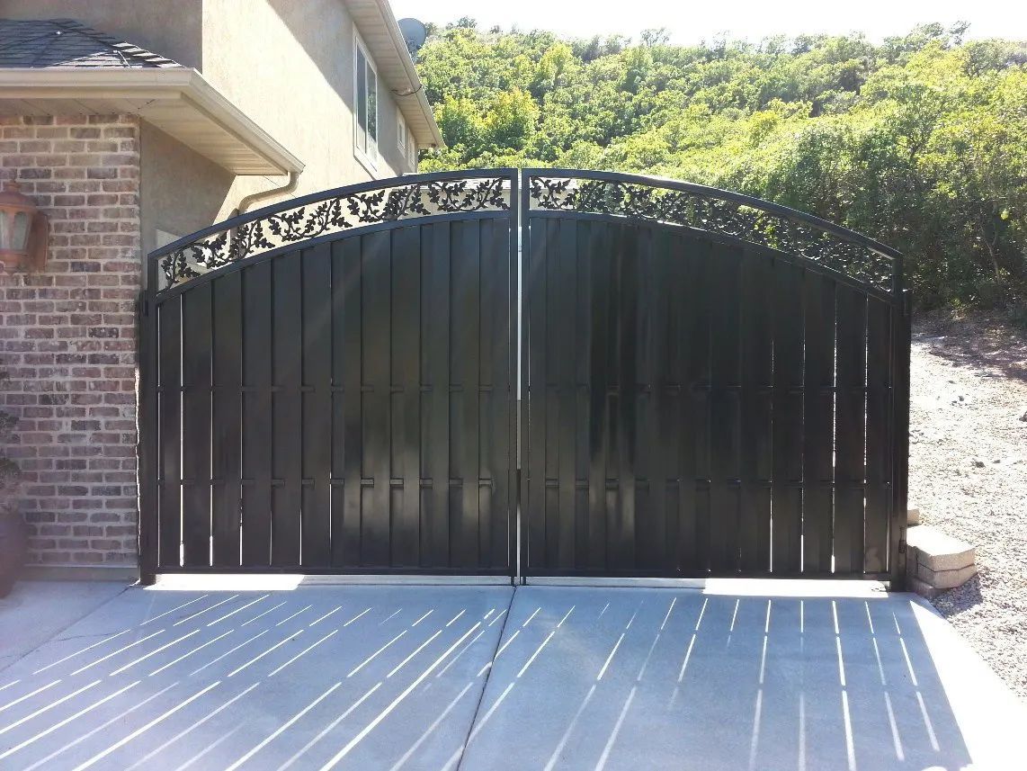 Black wrought-iron driveway gate in front of a brick house with sunlit concrete driveway