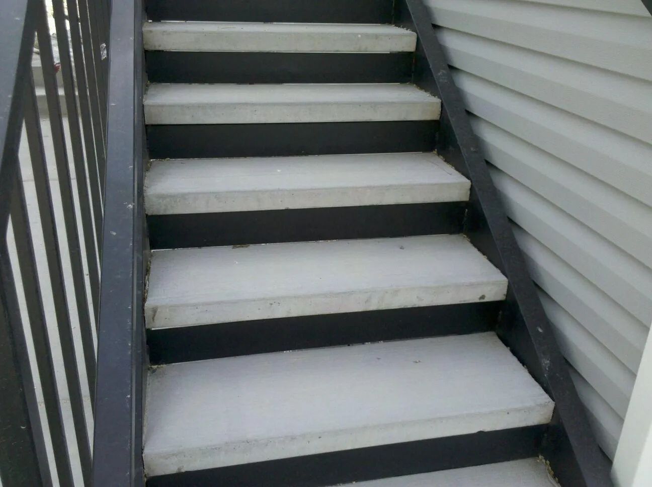 Snow-covered outdoor stairs beside gray siding and a black railing
