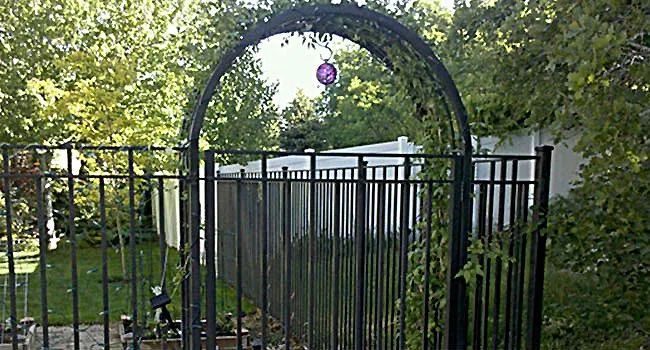Garden gate with black metal fence and arched trellis surrounded by green foliage and a purple hanging ornament