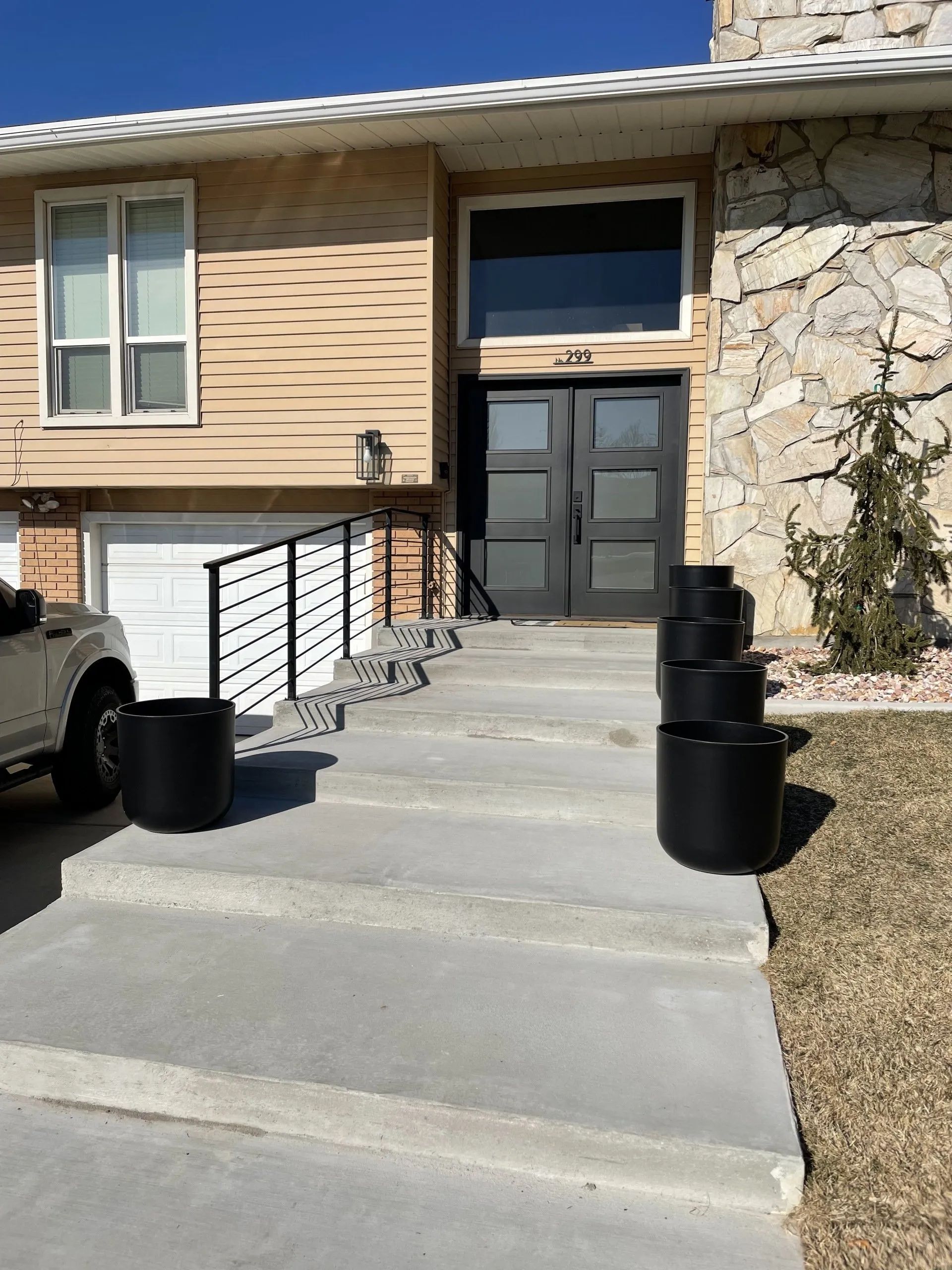 Front entrance of a tan house with black double doors, stone wall, and concrete walkway with railings and planters
