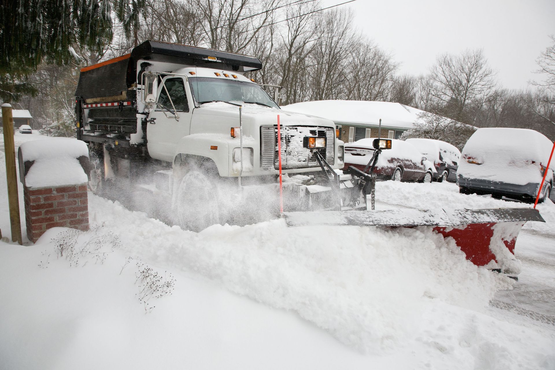 Black pickup truck with snowplow clearing snow in front of a white garage during a snowstorm.