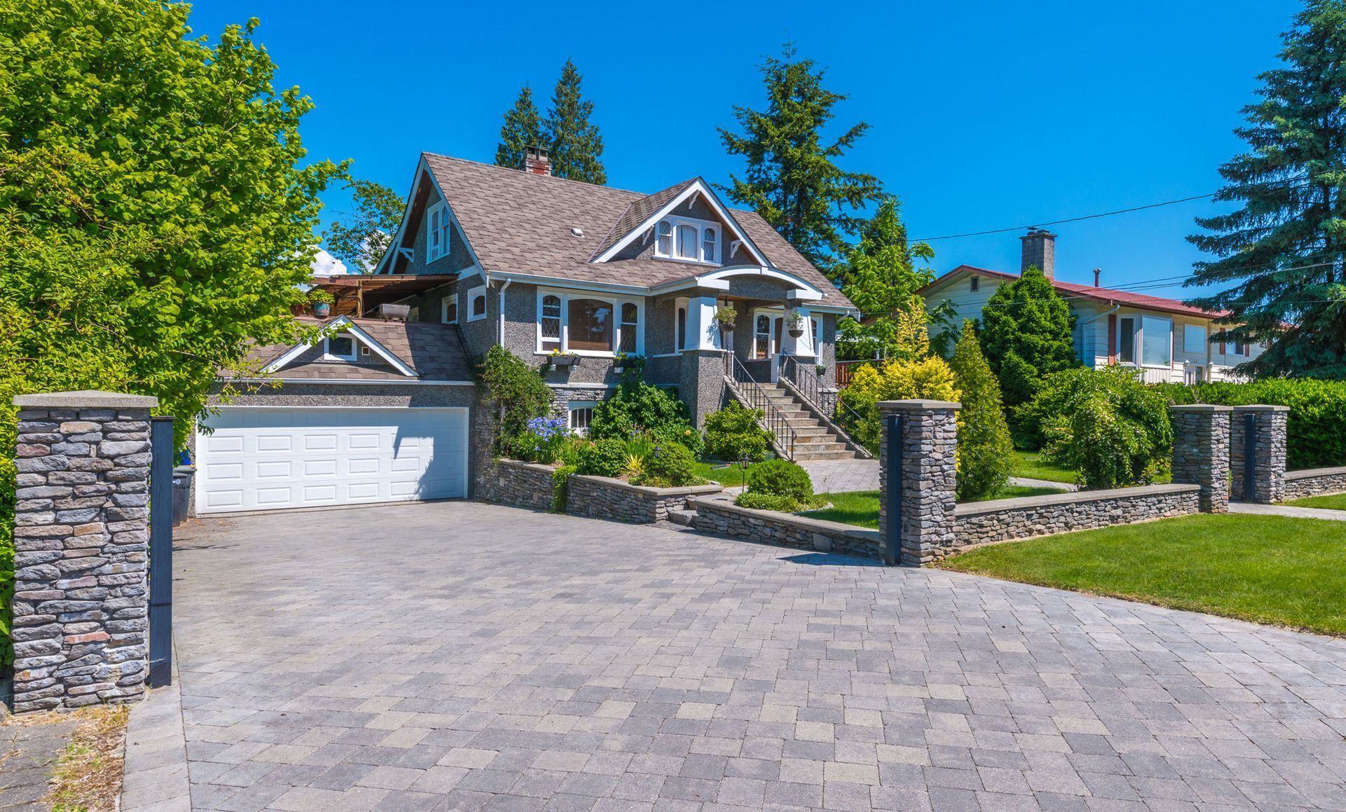 Stone-fronted house with gray roof, brick pillars, driveway, and lawn on a sunny day.