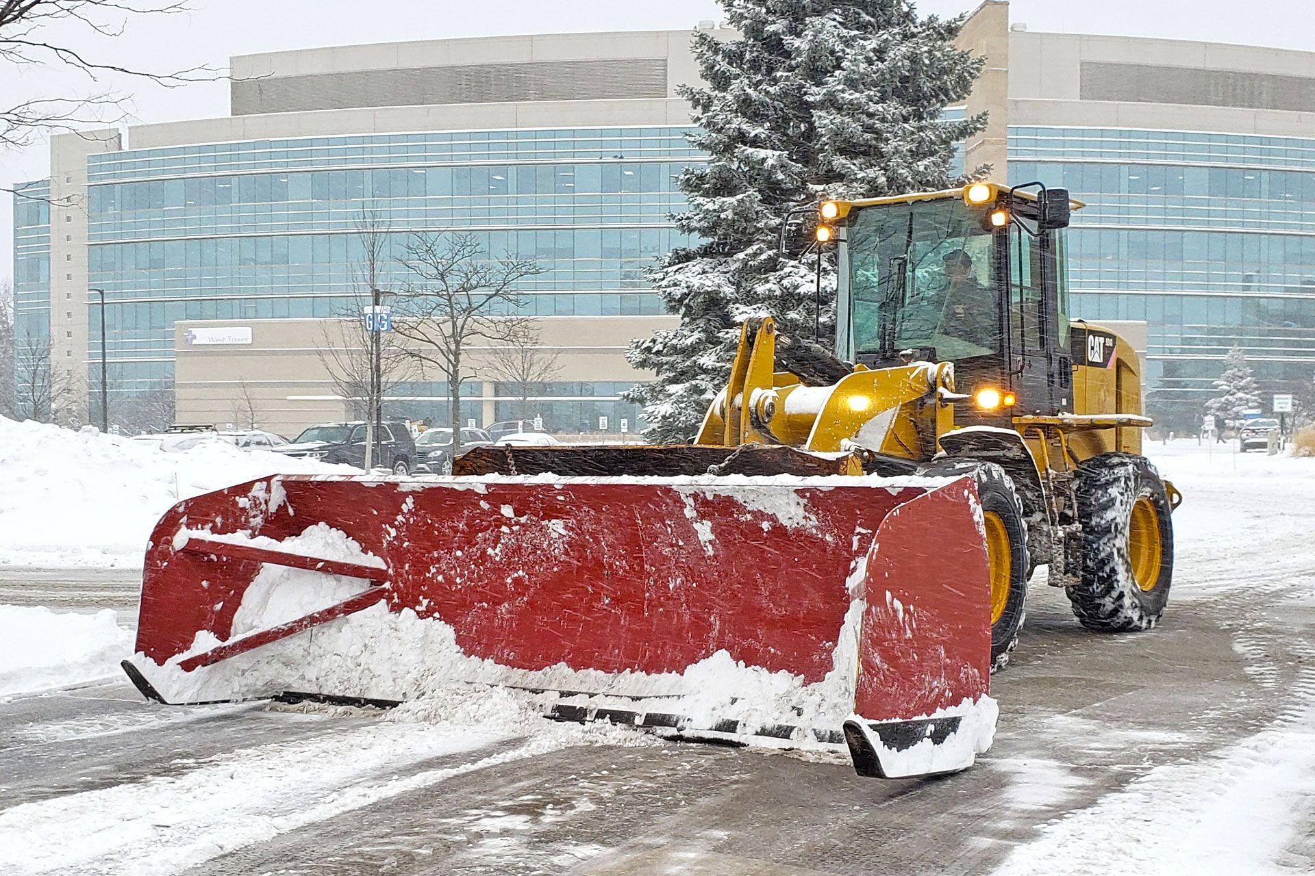 Snowplow clearing a road in front of a modern building during a snowstorm.