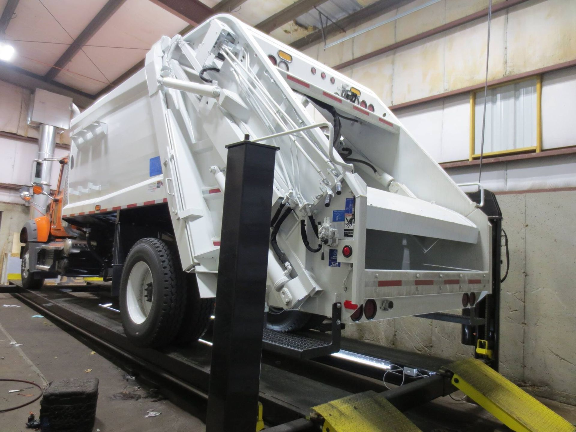 White garbage truck raised on a lift inside a building, showing the back and side.
