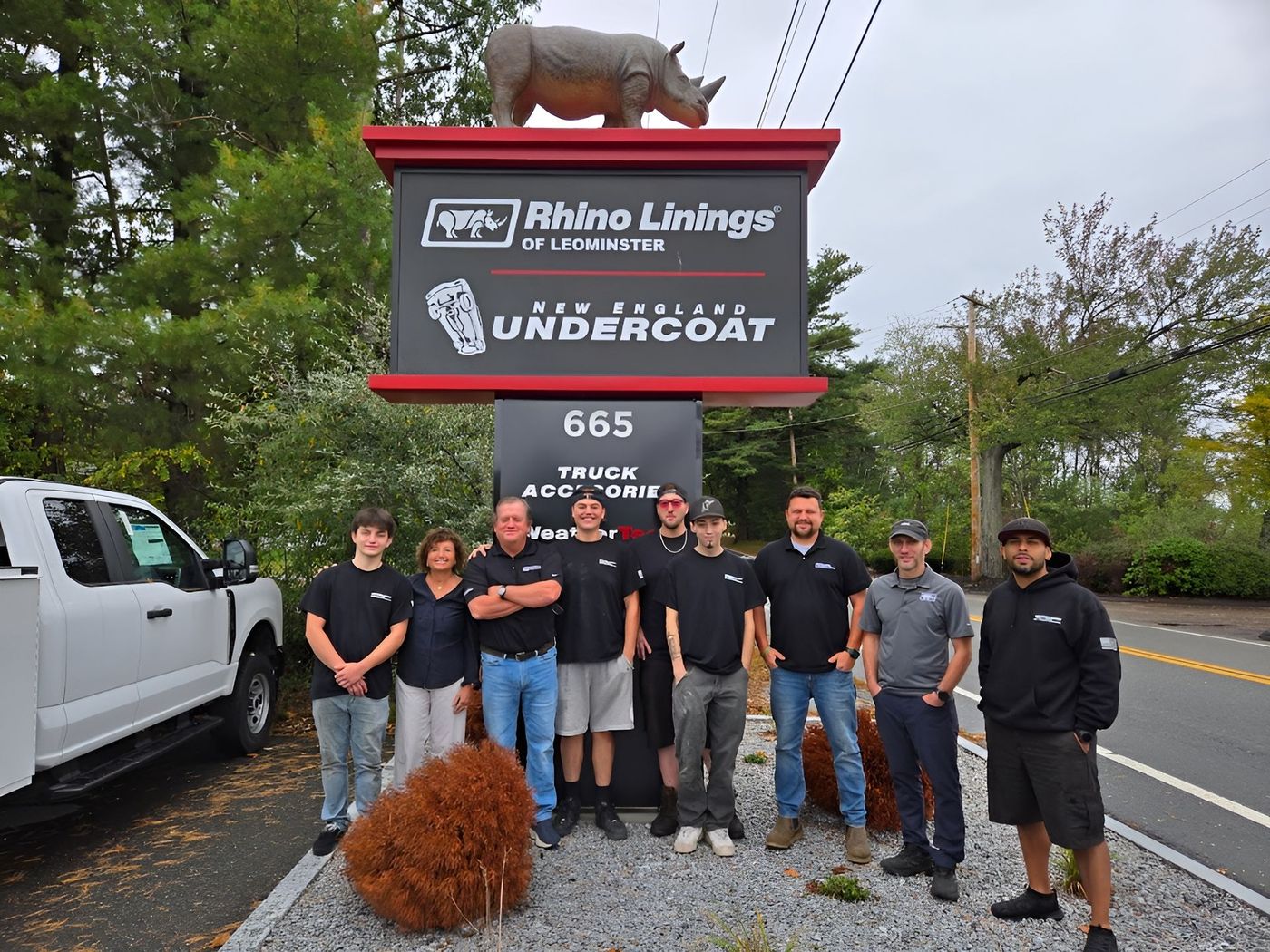 Group of people standing in front of a Rhino Linings sign, a truck parked nearby.