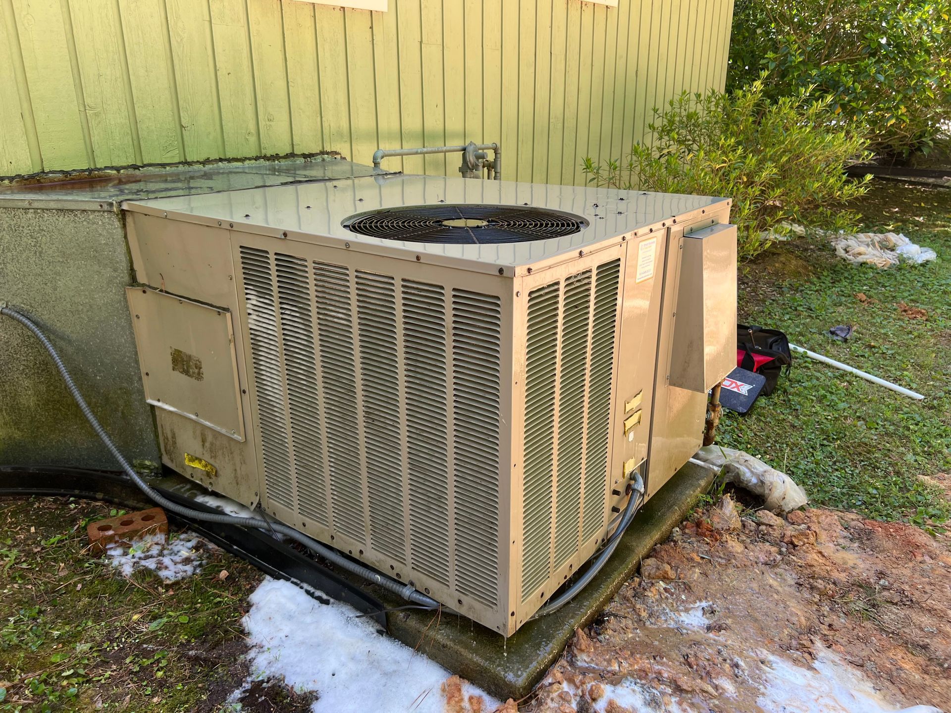 Outdoor air conditioning unit beside a house, with snow on the ground and a garden hose nearby - after