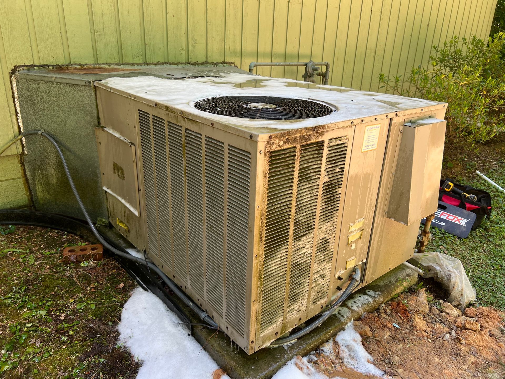 Outdoor air conditioning unit beside a house, with snow on the ground and a garden hose nearby - before