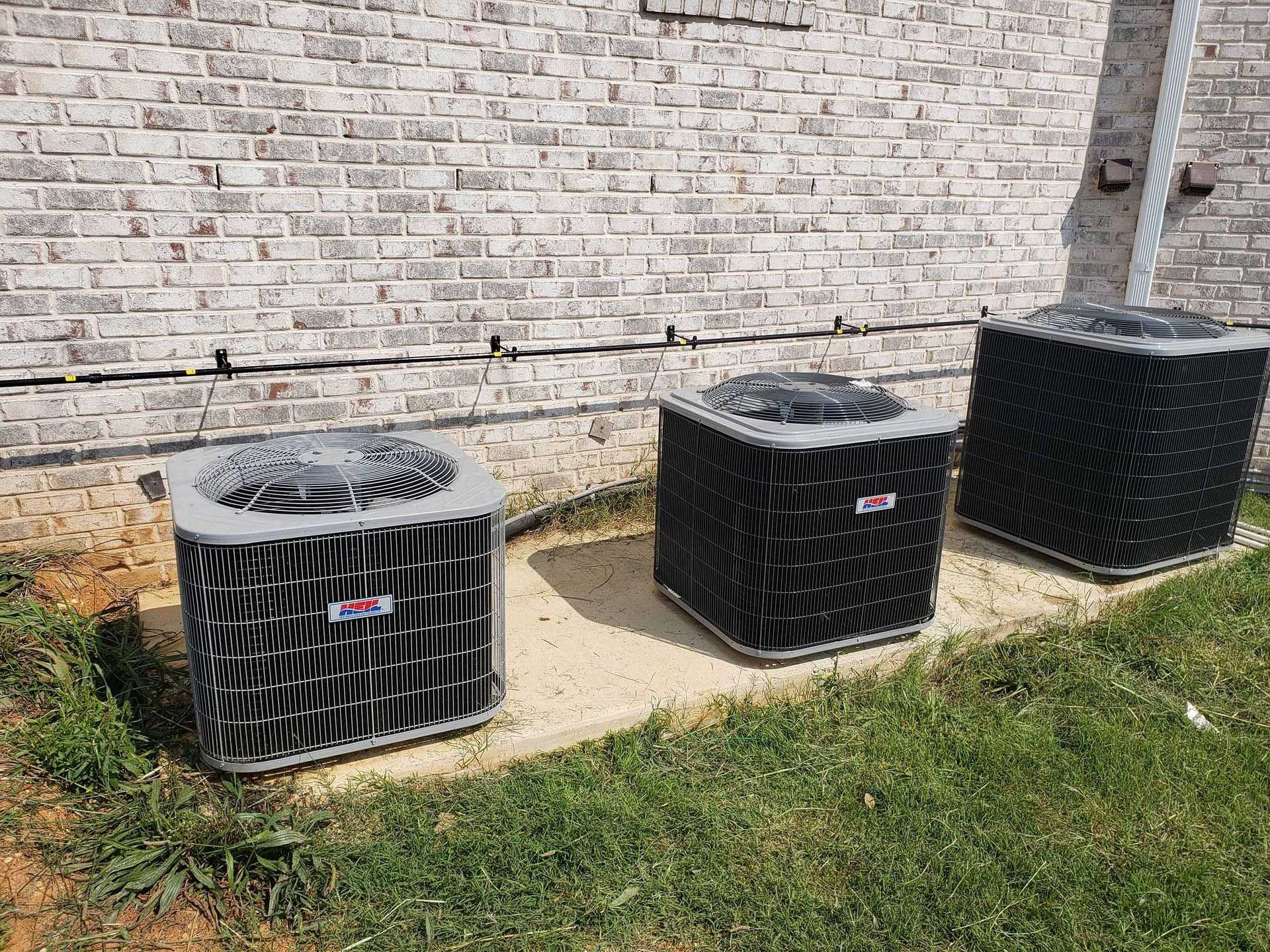 Three air conditioning units outside, set on concrete pads, near a brick building.