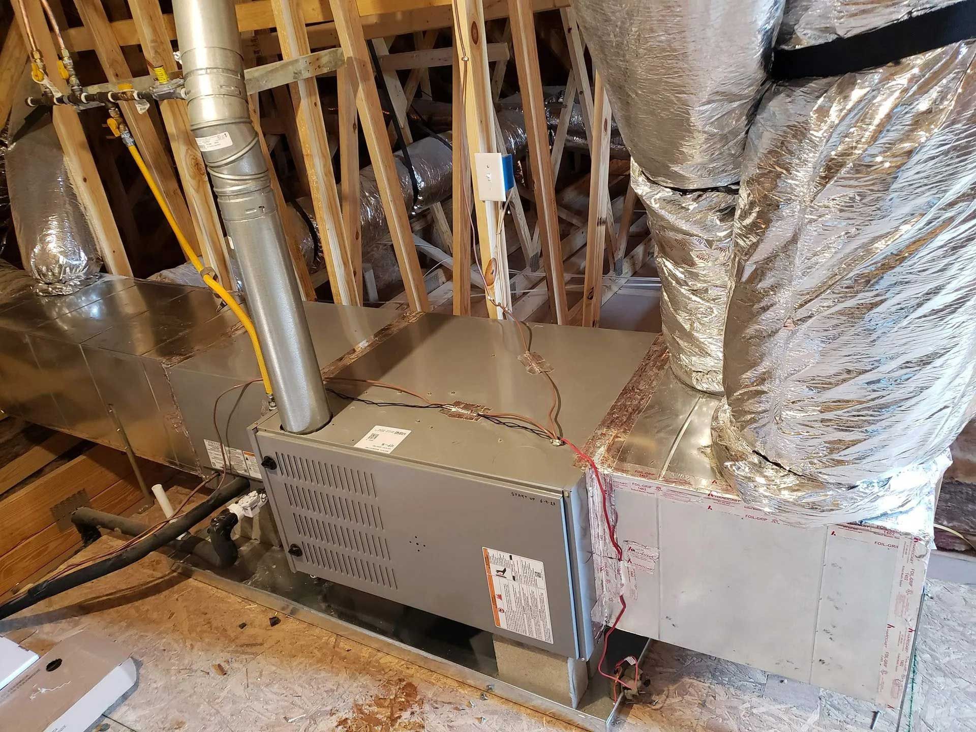 HVAC system in an attic. Metallic furnace and ductwork visible. Wooden beams in background.