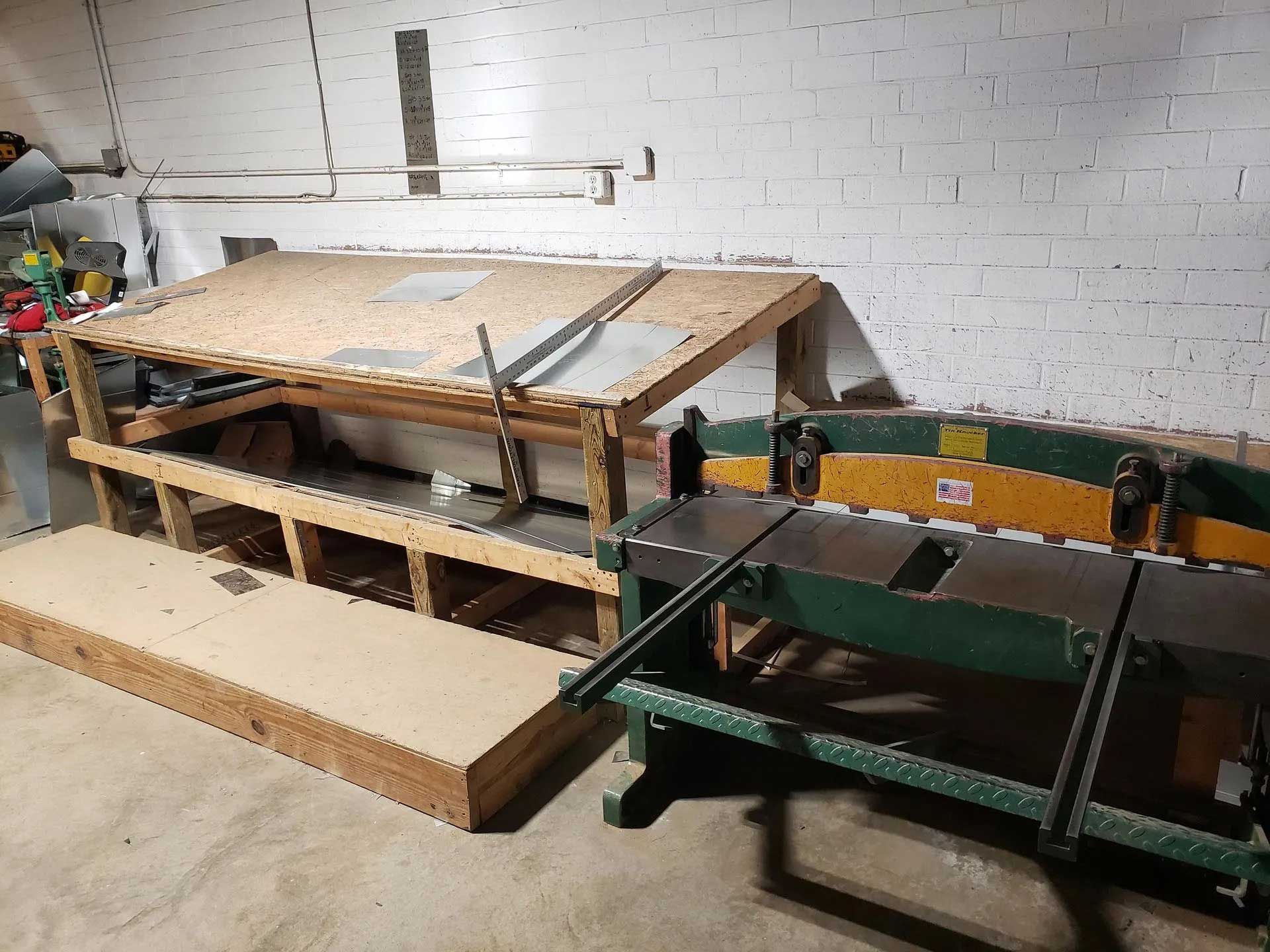 A workshop with a wooden workbench, a large sheet metal shear, and a wood plank.