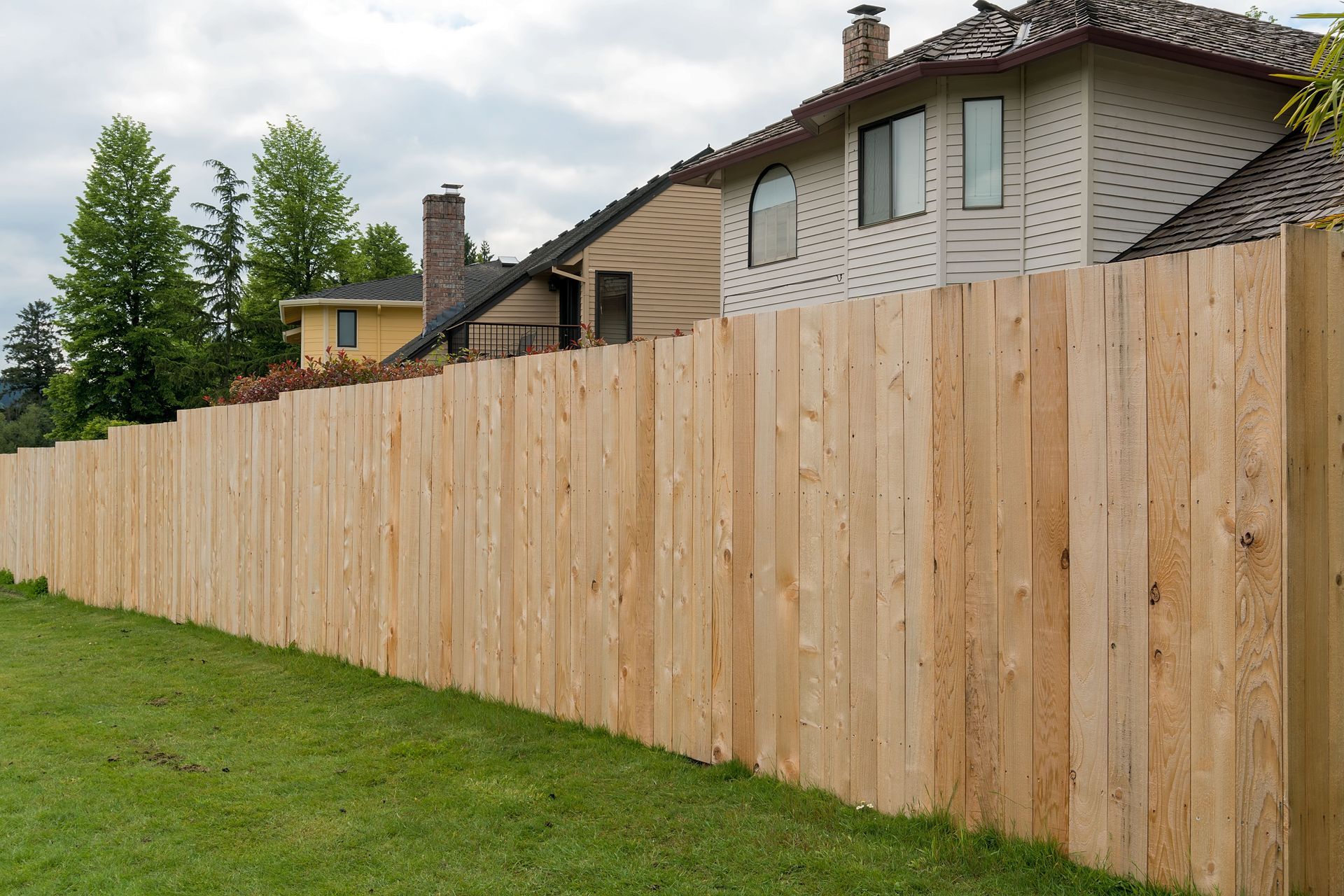 A new, light-colored wooden privacy fence stands in front of residential houses on a green lawn under a cloudy sky.