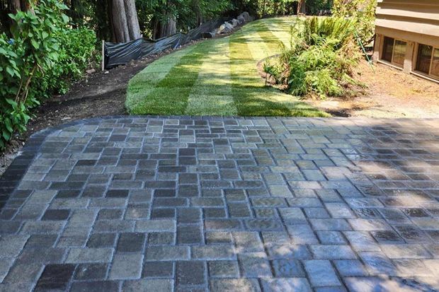 A paved patio with a dark grey stone pattern sits adjacent to a newly installed green lawn and a house exterior.