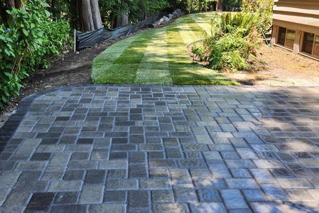 A paved patio with a dark grey stone pattern sits adjacent to a newly installed green lawn and a house exterior.