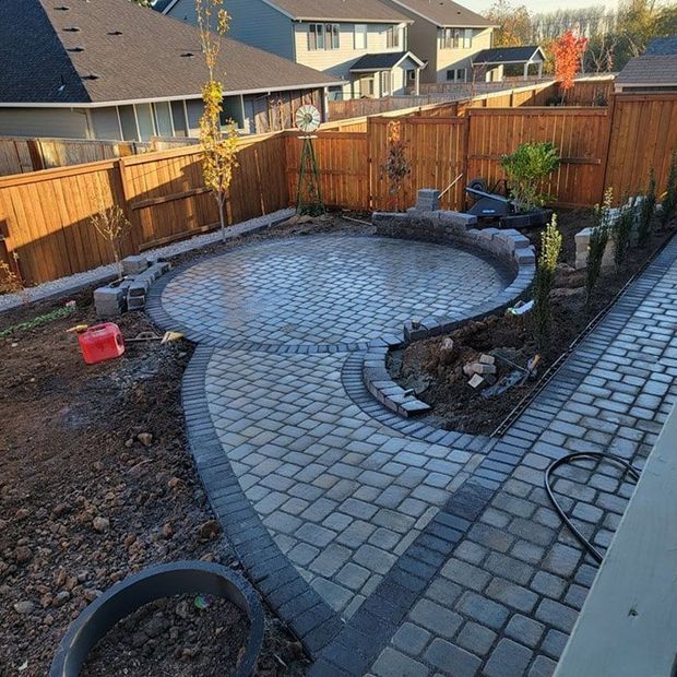 A newly installed stone paver patio and walkway in a backyard, bordered by a wooden fence with landscaping still in progress.