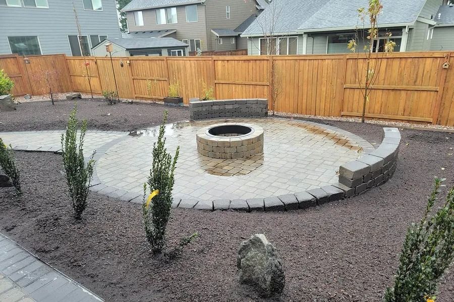 A circular paver patio with a stone fire pit and curved seating wall, surrounded by dark gravel and small shrubs.