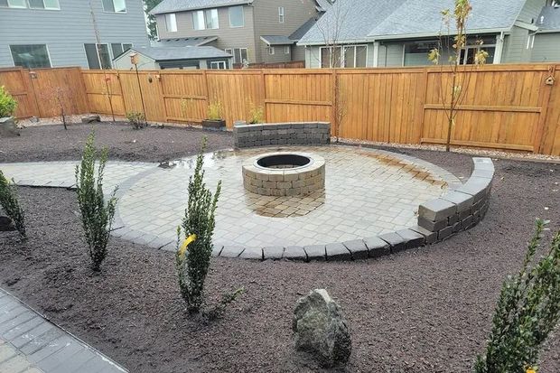 A circular stone patio with a fire pit and curved seating wall, surrounded by dark landscaping mulch and young shrubs.