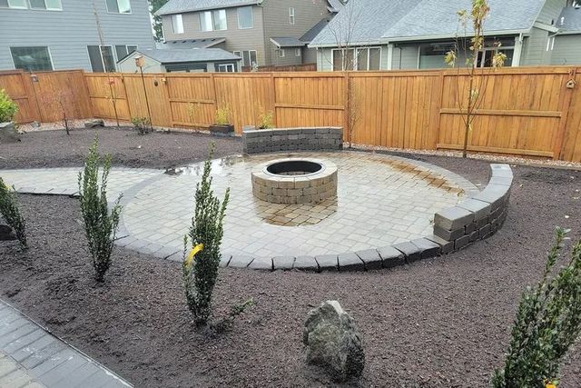 A circular stone patio with a fire pit and curved seating wall, surrounded by dark landscaping mulch and young shrubs.