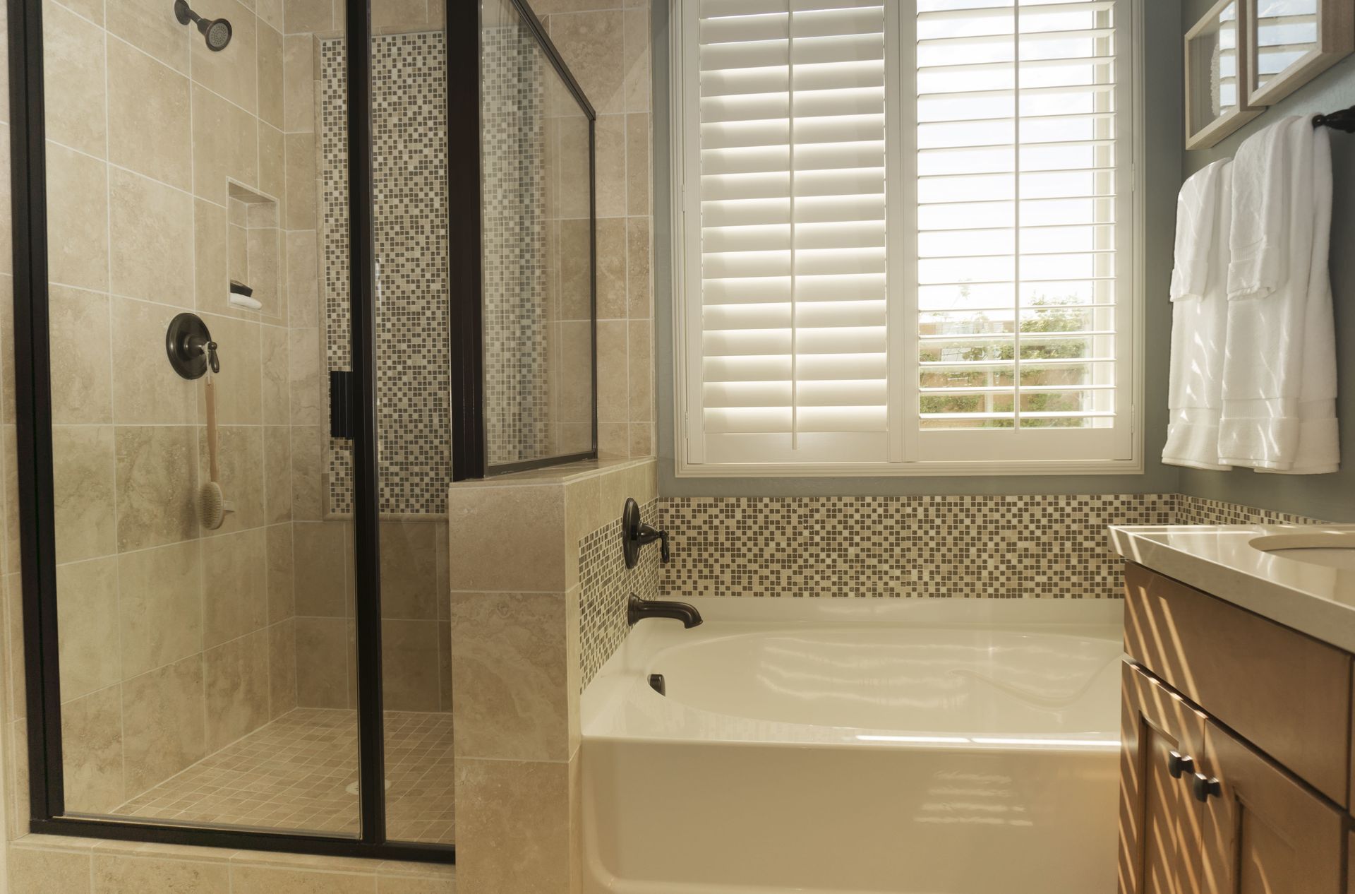 Bathroom with a glass shower, white bathtub, and wood cabinets. Light-colored tile and shutters.