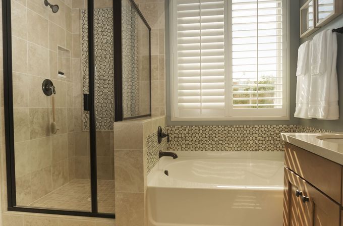 Bathroom with a glass shower, white bathtub, and wood cabinets. Light-colored tile and shutters.