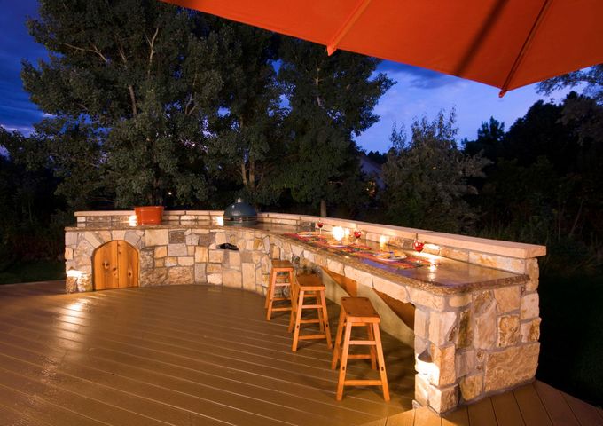 Outdoor stone bar with stools, grill, and oven on a wooden deck at dusk. Orange umbrella overhead.