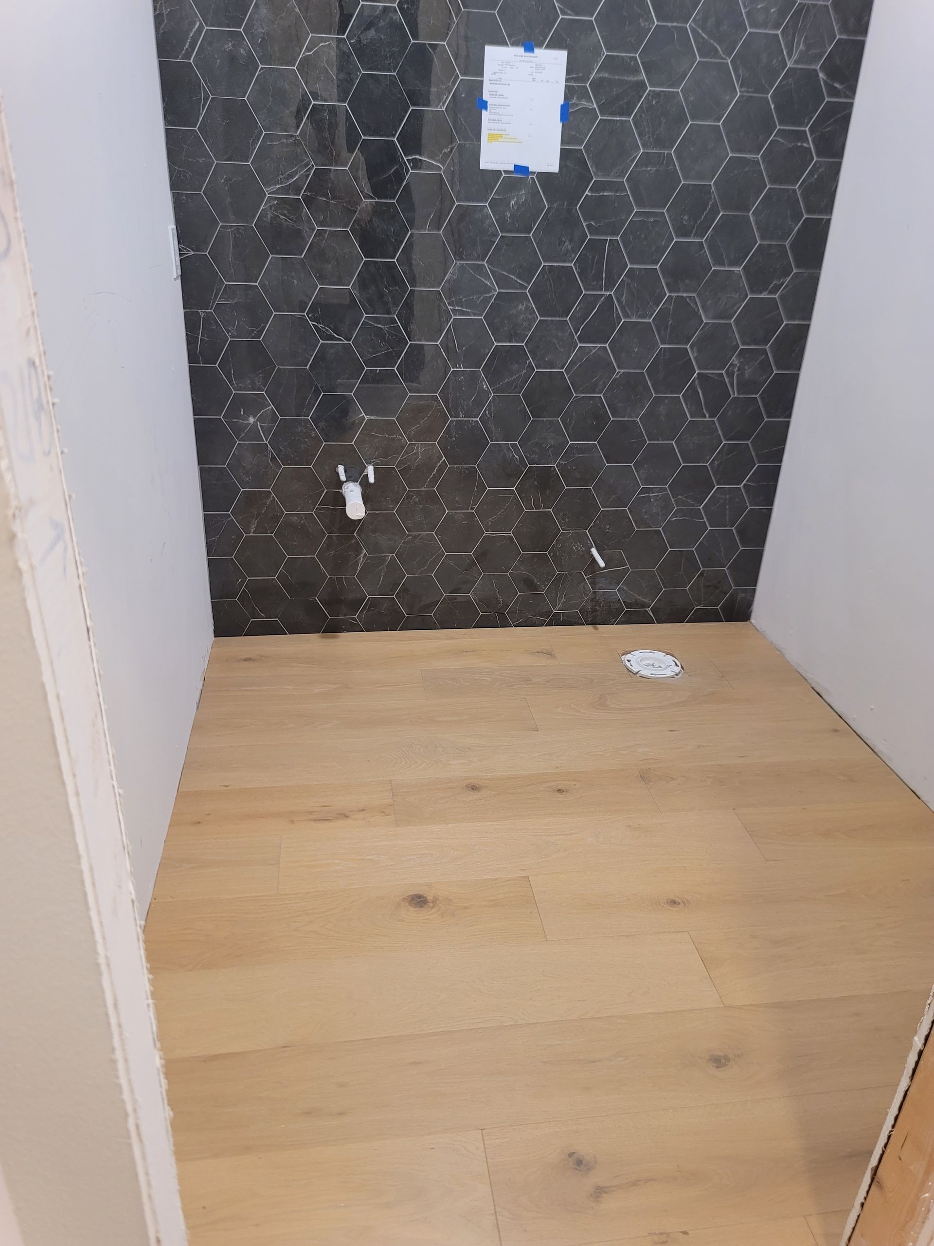Interior view of a small room. Dark hexagonal tile wall, light wood floor, white walls.