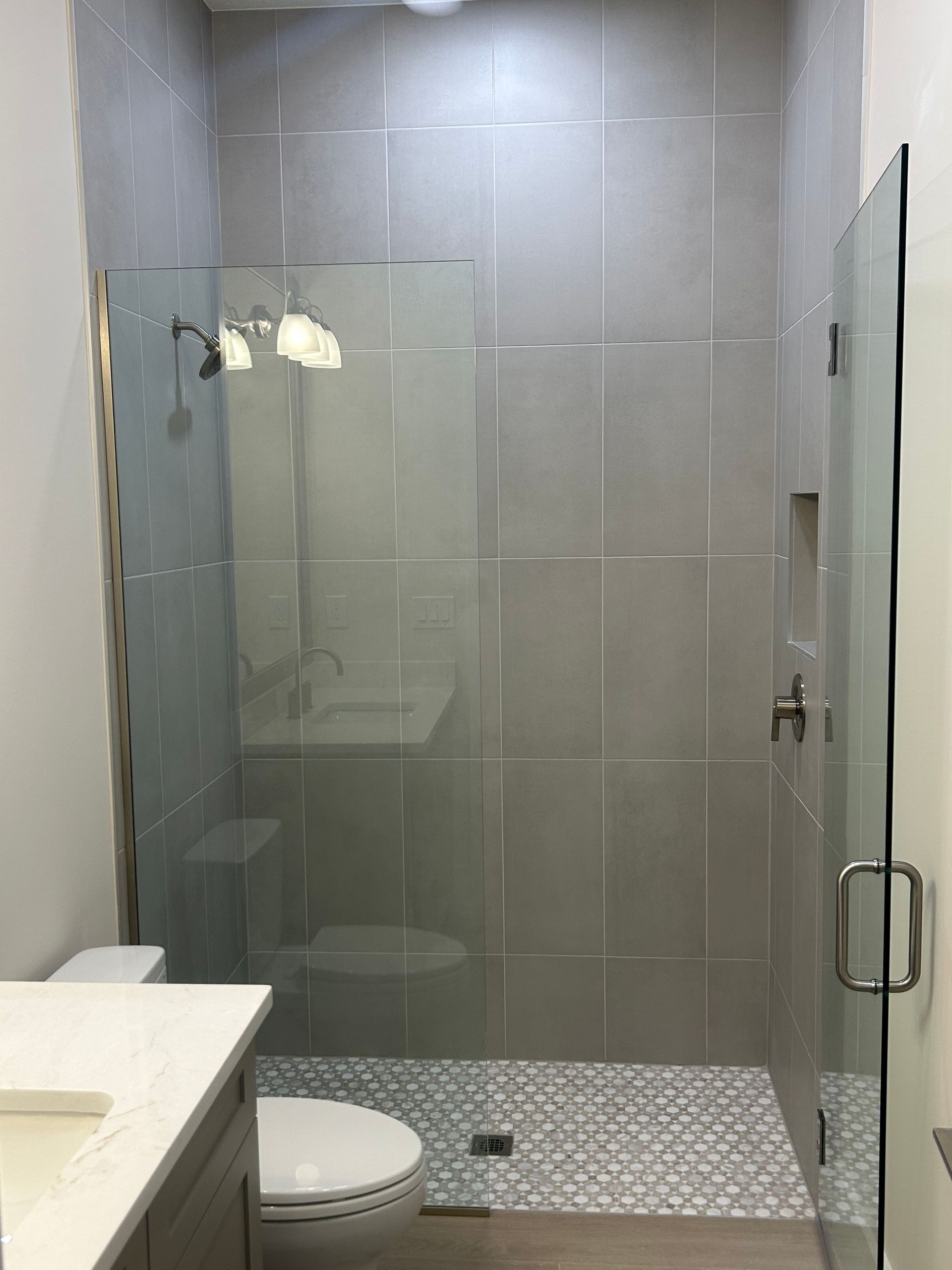 Modern bathroom with gray tiled walls, glass shower, and mosaic floor.