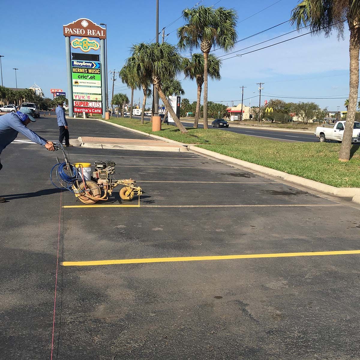 Worker moving a yellow pavement-stripping machine in a parking lot near a roadside sign and palm trees