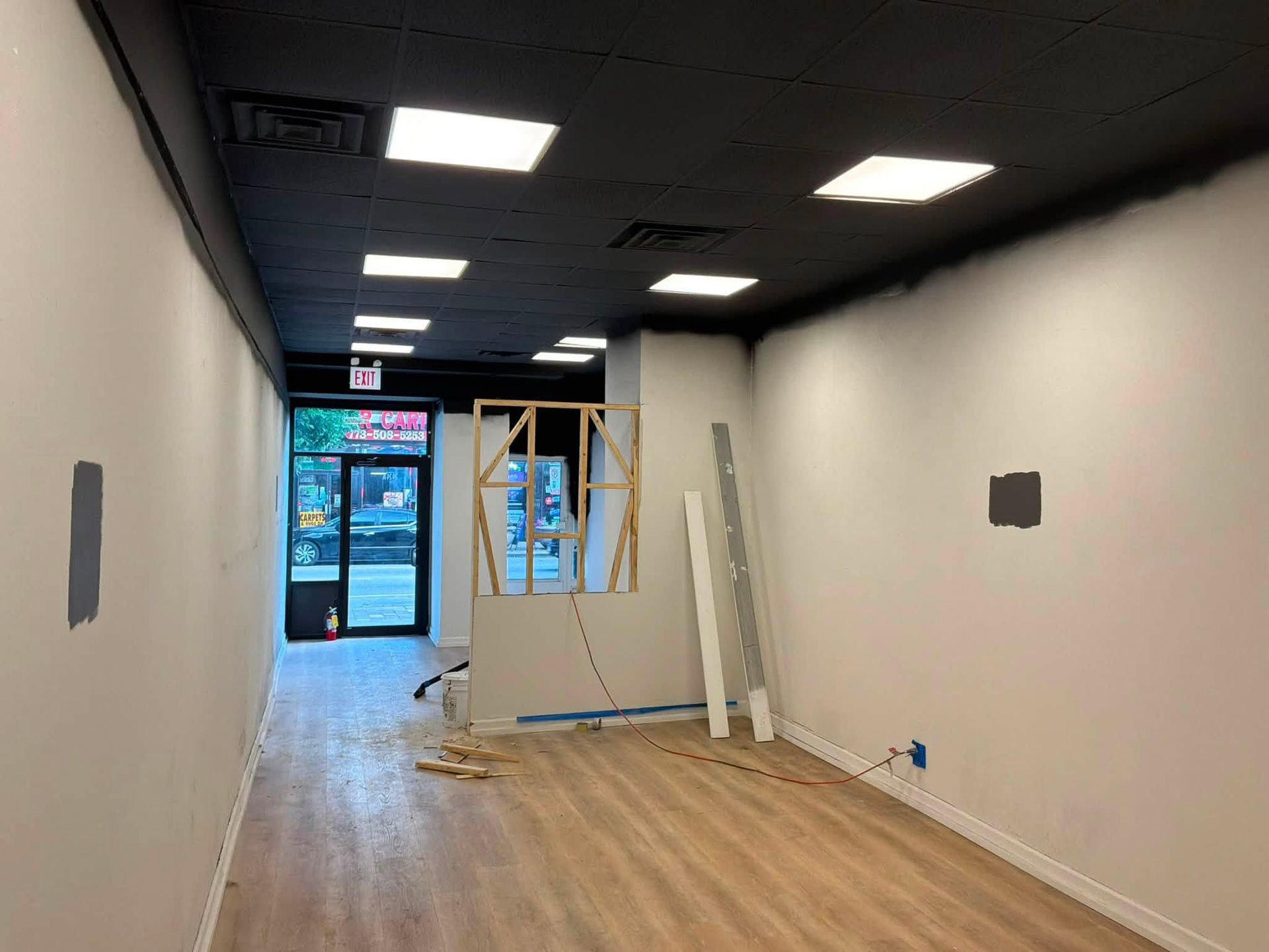 Empty office hallway with white walls, black ceiling, wood floor, and an open doorway at the end under renovation.