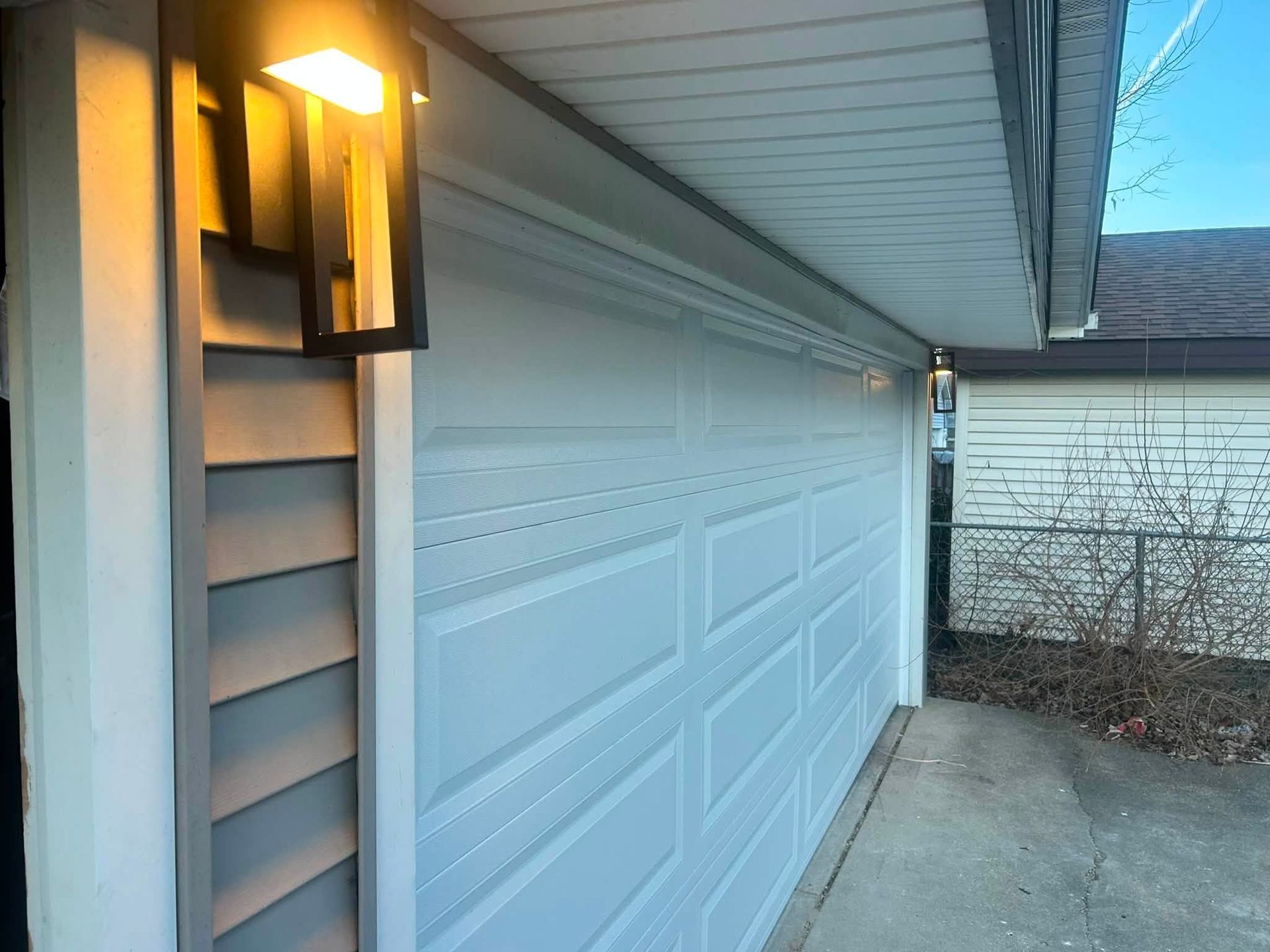 White garage door beside house siding with lit wall lantern and concrete driveway at dusk