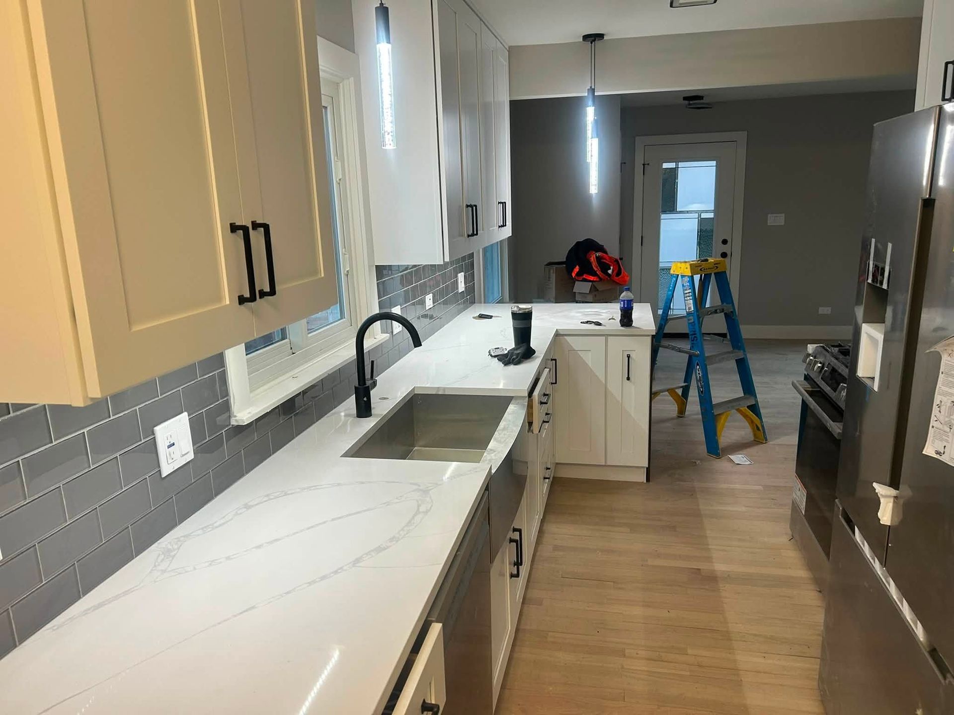 Modern kitchen under renovation with white cabinets, sink, tile backsplash, and a ladder in the hall.