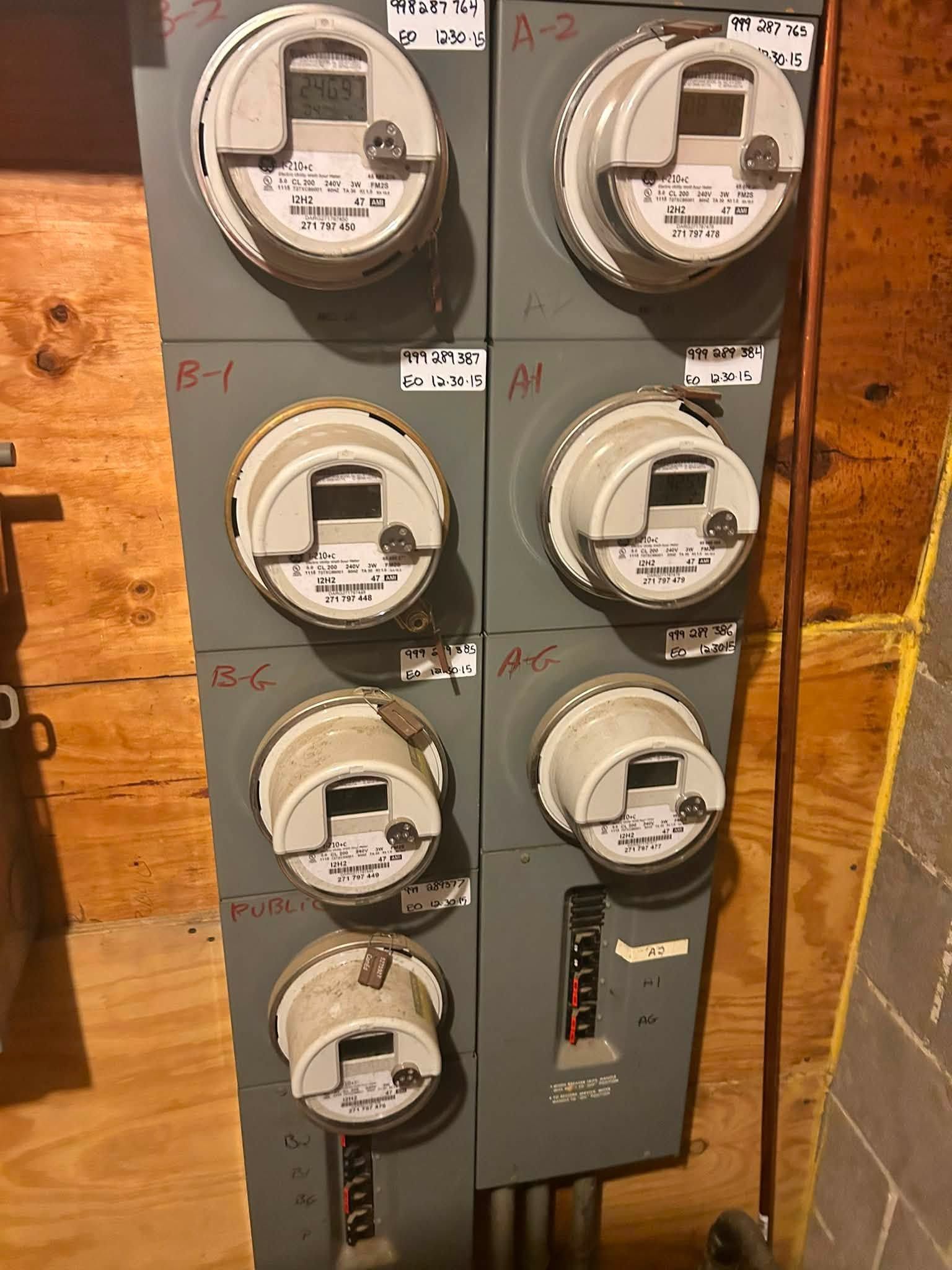 A row of electric meters mounted on a wooden wall panel, with gray meter boxes and circular glass dials.