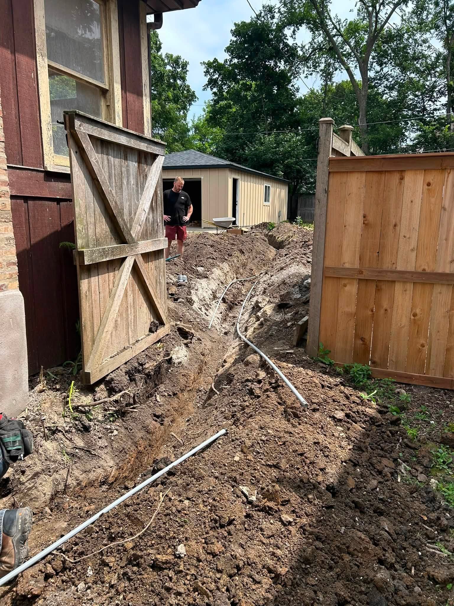Muddy trench beside a house with an open wooden gate and fence, with a person in the background