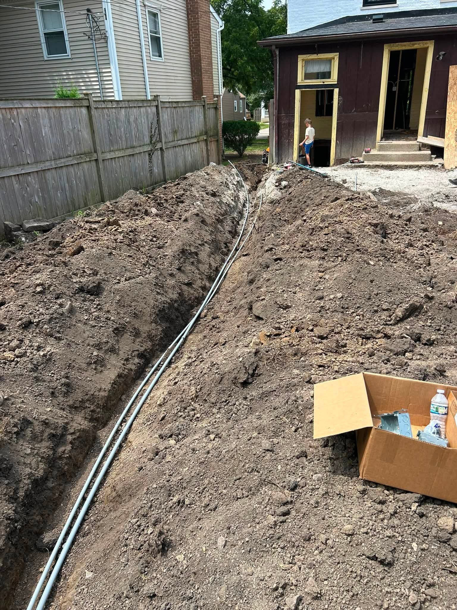 Narrow muddy trench beside a house, with pipes laid along the ground and a cardboard box in the foreground.