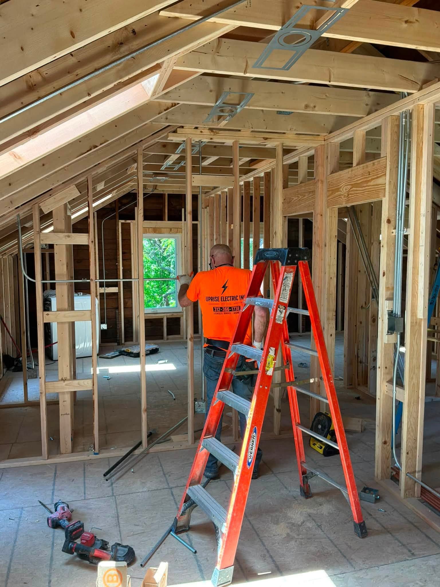 Interior of a house under construction, with red ladders, exposed framing, and a worker in an orange shirt.