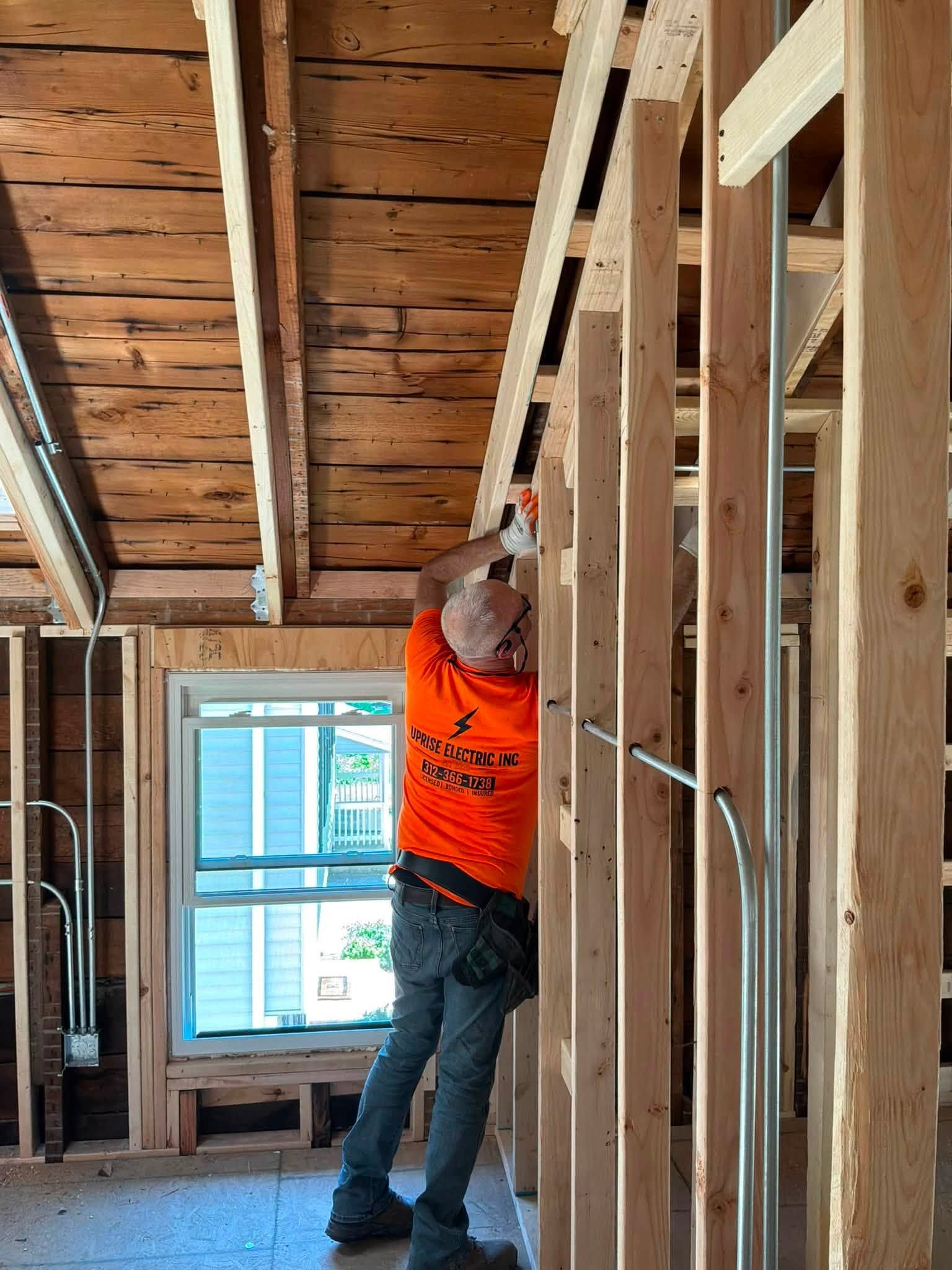 Construction worker in orange shirt installing drywall in an unfinished room with exposed studs.
