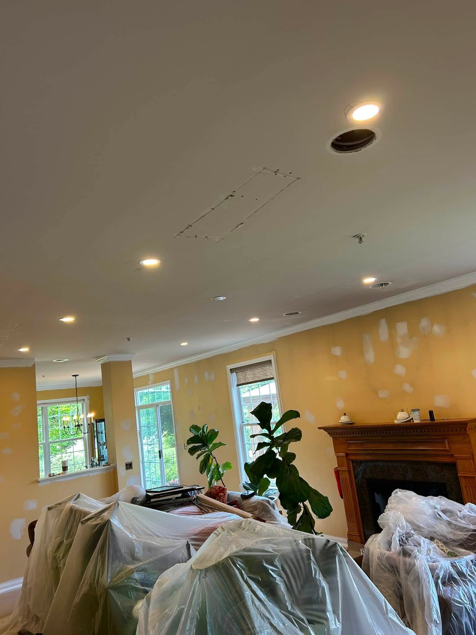 Living room with plastic-covered furniture, yellow walls, recessed lights, windows, and a fireplace under renovation