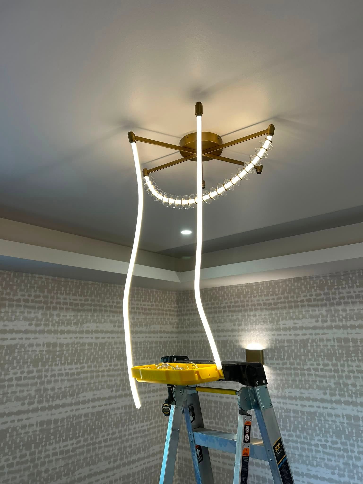 Ceiling light fixture hanging from the ceiling with a ladder below during installation, lit in a room.