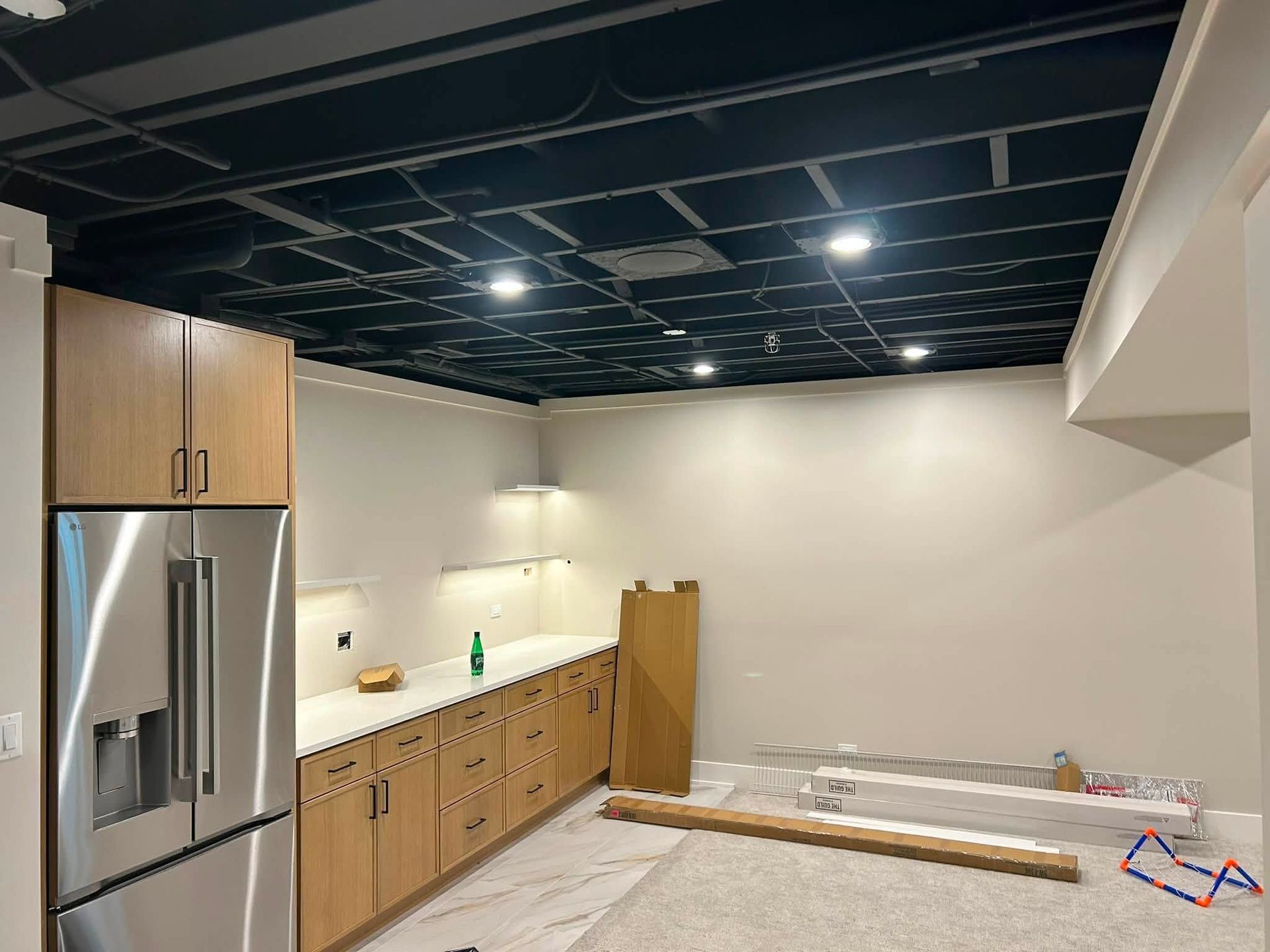 Unfinished kitchen with stainless fridge, wood cabinets, white walls, and exposed black ceiling.