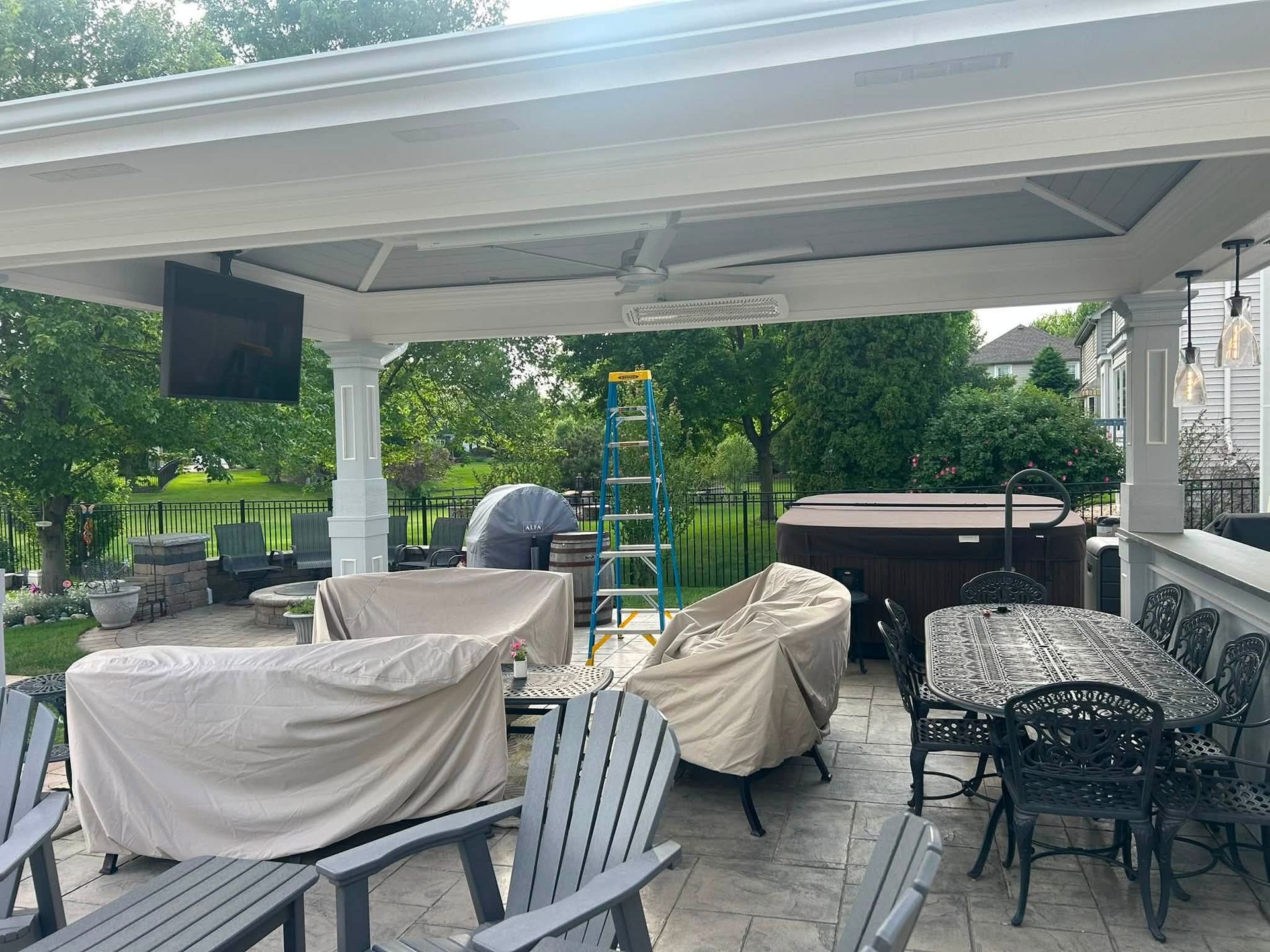 Covered patio with outdoor furniture under a white pavilion, surrounded by green trees and a garden.