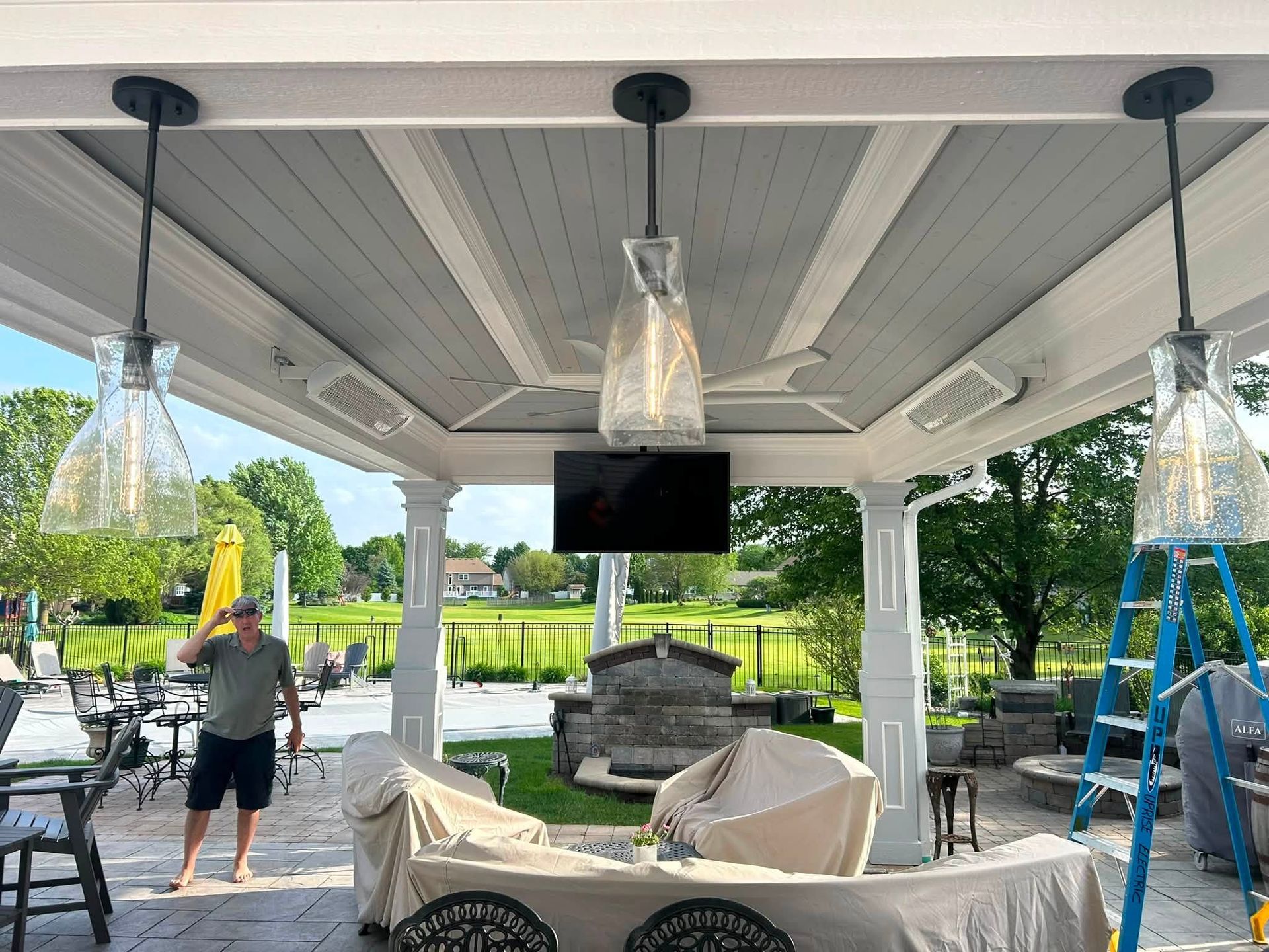 Covered patio with hanging lanterns, TV, seating, and a person on a ladder near blue trim repairs