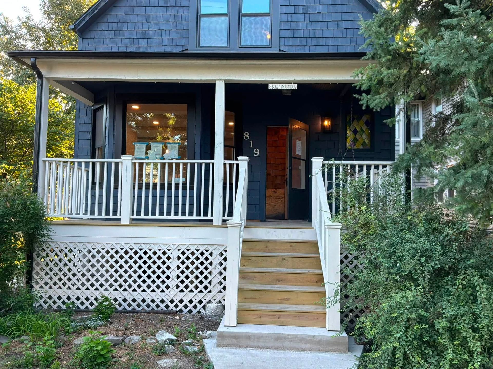 Front porch of a dark house with white railings, steps, and lattice skirting, surrounded by greenery.